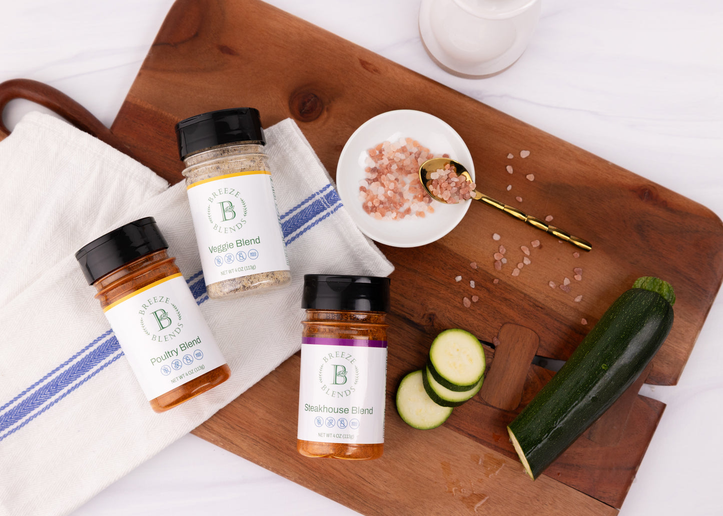 Breeze Blends BBQ pack of three jars on a wooden cutting board with zucchini and a small bowl of pink salt.