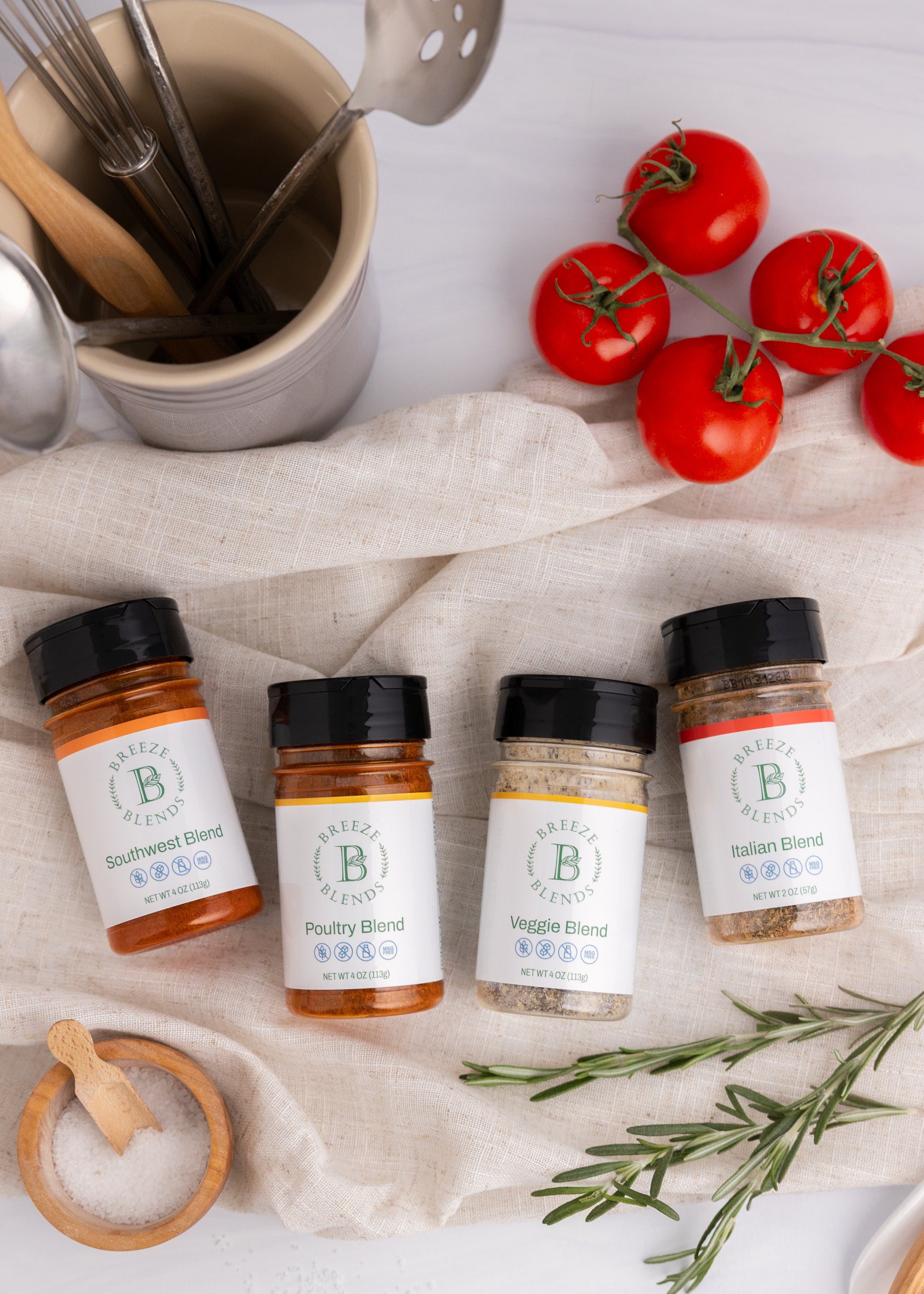 Collection of Breeze Blends spice jars with labels on a tablecloth with tomatoes and rosemary