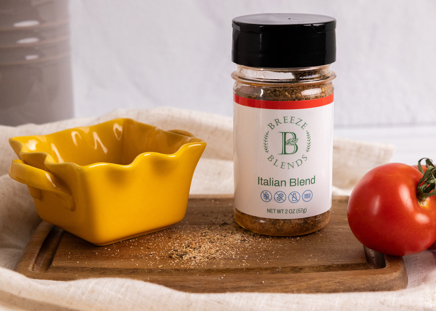 Bottle of Breeze Blends Italian Blend seasoning on a wooden cutting board with a yellow dish and tomato.